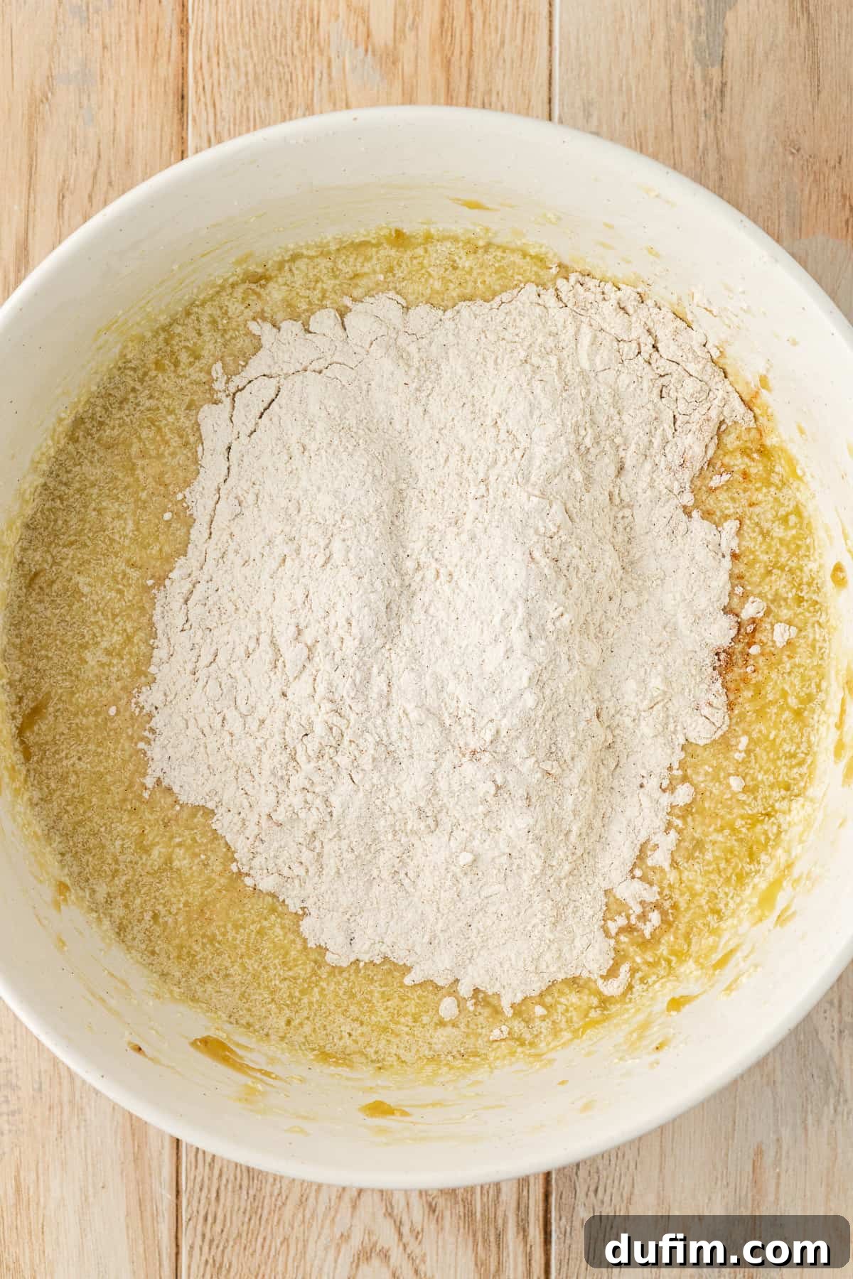 A white bowl on a wood surface showing wet ingredients for banana cake with the dry flour mixture sitting on top, yet to be mixed in, illustrating the combining step