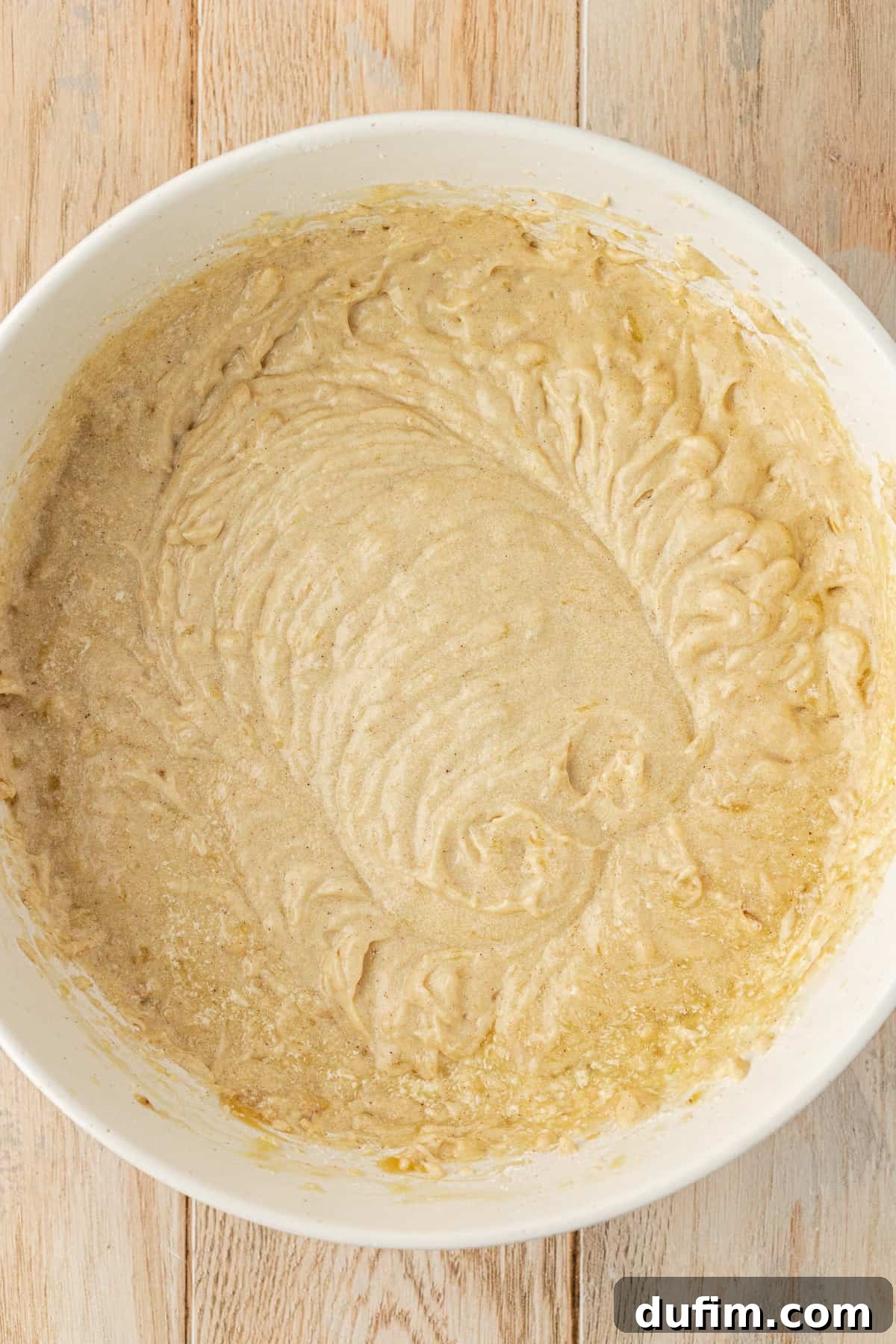A white bowl sitting on a wood surface, filled with smooth, ready-to-bake banana cake batter