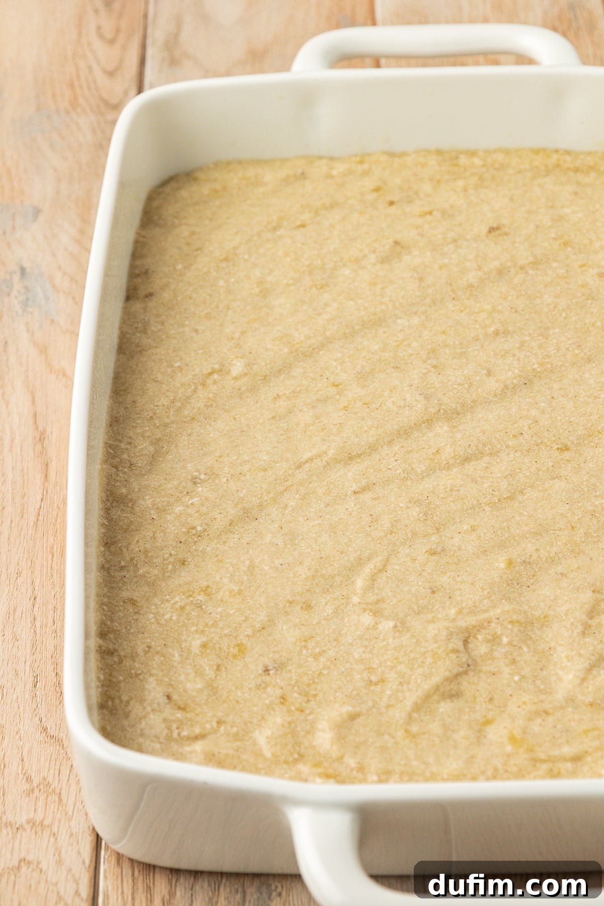 Banana cake batter evenly spread in a white 9x13 baking dish on a wood surface, poised for baking