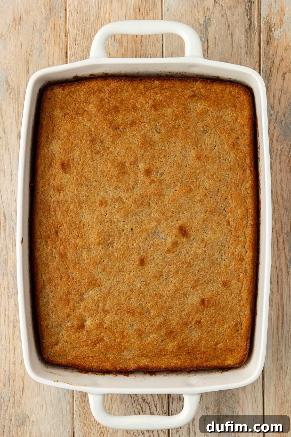 Freshly baked golden-brown banana cake in a 9x13 white dish, resting on a wood surface, cooling down before frosting