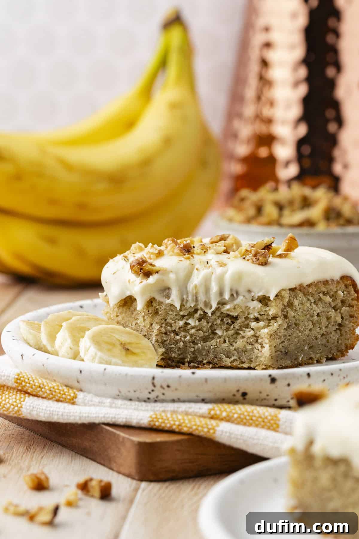 A perfect slice of banana cake on a small plate, resting on a white and yellow striped towel on a wooden board, surrounded by the remaining cake, a bowl of walnuts, and whole bananas, ready to be enjoyed