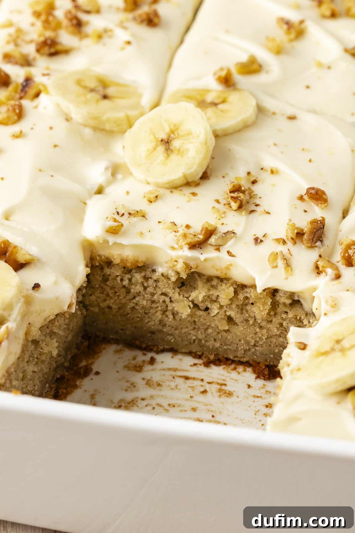 Close up of a white baking dish filled with moist banana cake topped with creamy cream cheese frosting, with one slice missing revealing the distinct layers of cake and frosting