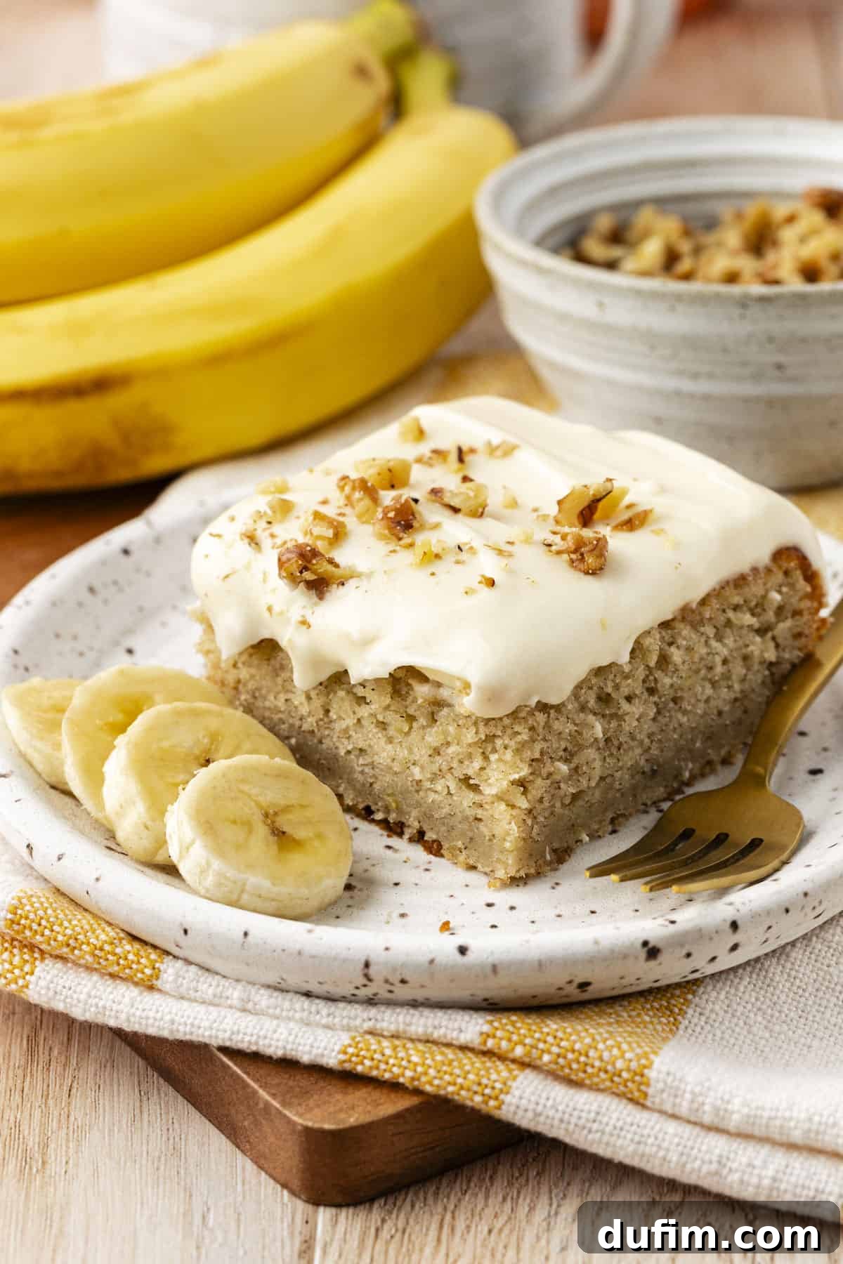 A delectable slice of banana cake with one bite missing, presented on a small plate alongside fresh banana slices, all arranged on a yellow and white striped kitchen towel with whole bananas and a bowl of crumb topping in the soft-focused background