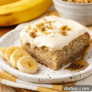 A slice of banana cake with one bite missing on a small plate with fresh banana slices, on top of a yellow and white striped kitchen towel with whole bananas and bowl of crumb topping in the background