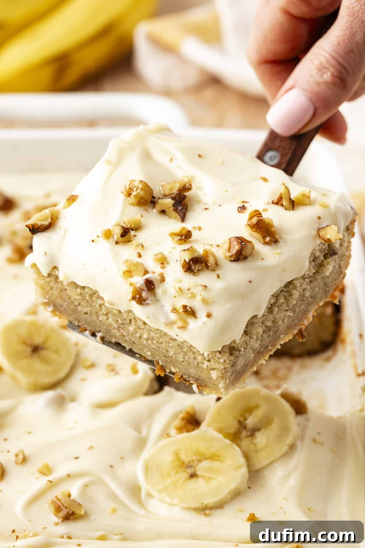 A slice of banana cake with chopped walnuts sprinkled on top, being lifted out of the cake dish with a spatula, highlighting its moist texture and generous frosting layer