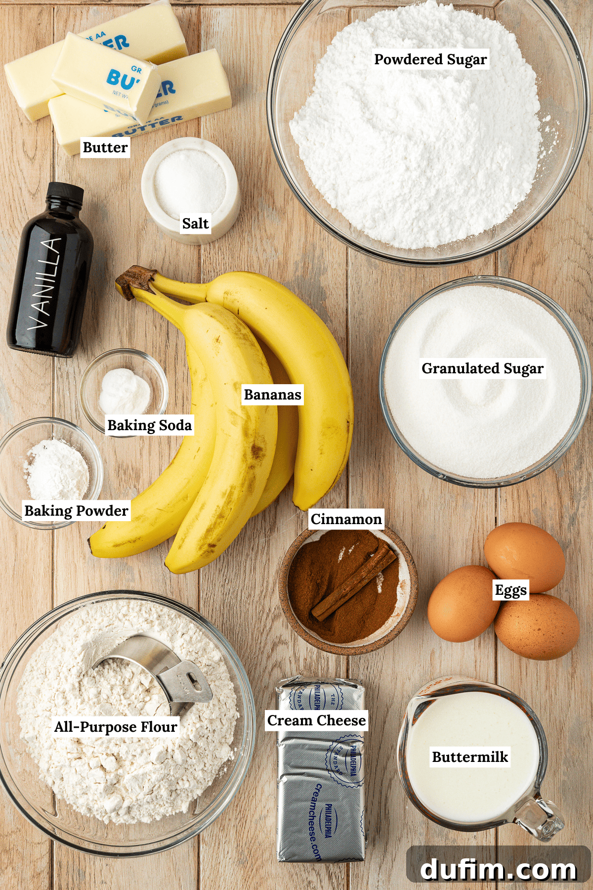 Overhead view of all the fresh ingredients for banana cake laid out on a wooden surface, including bowls of flour, sugar, butter, eggs, bananas, buttermilk, and cream cheese, ready for baking