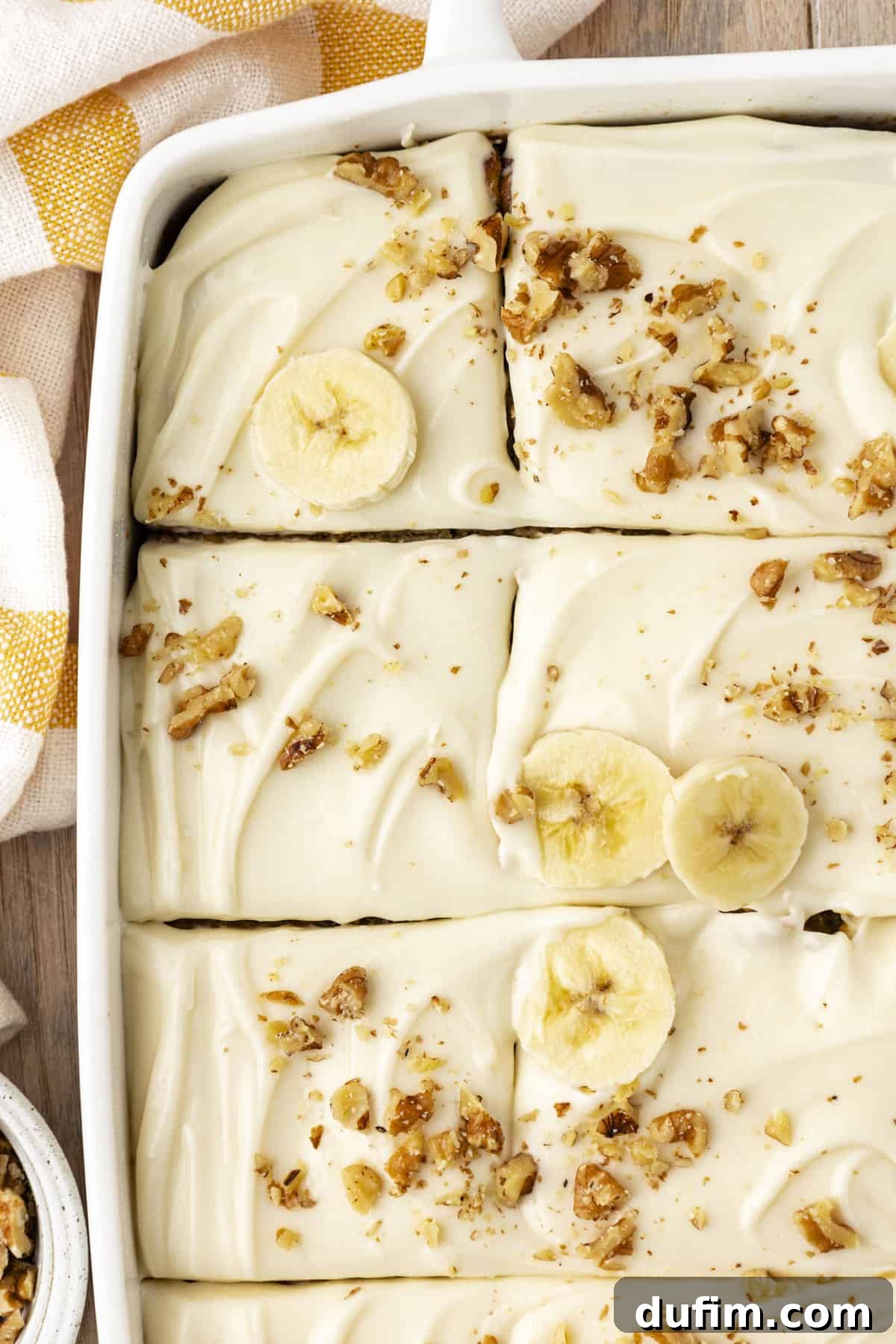 Overhead view of a baked banana cake in a white baking dish, beautifully sliced into pieces and garnished with fresh banana slices and chopped walnuts, with a charming yellow and white striped kitchen towel beside the pan