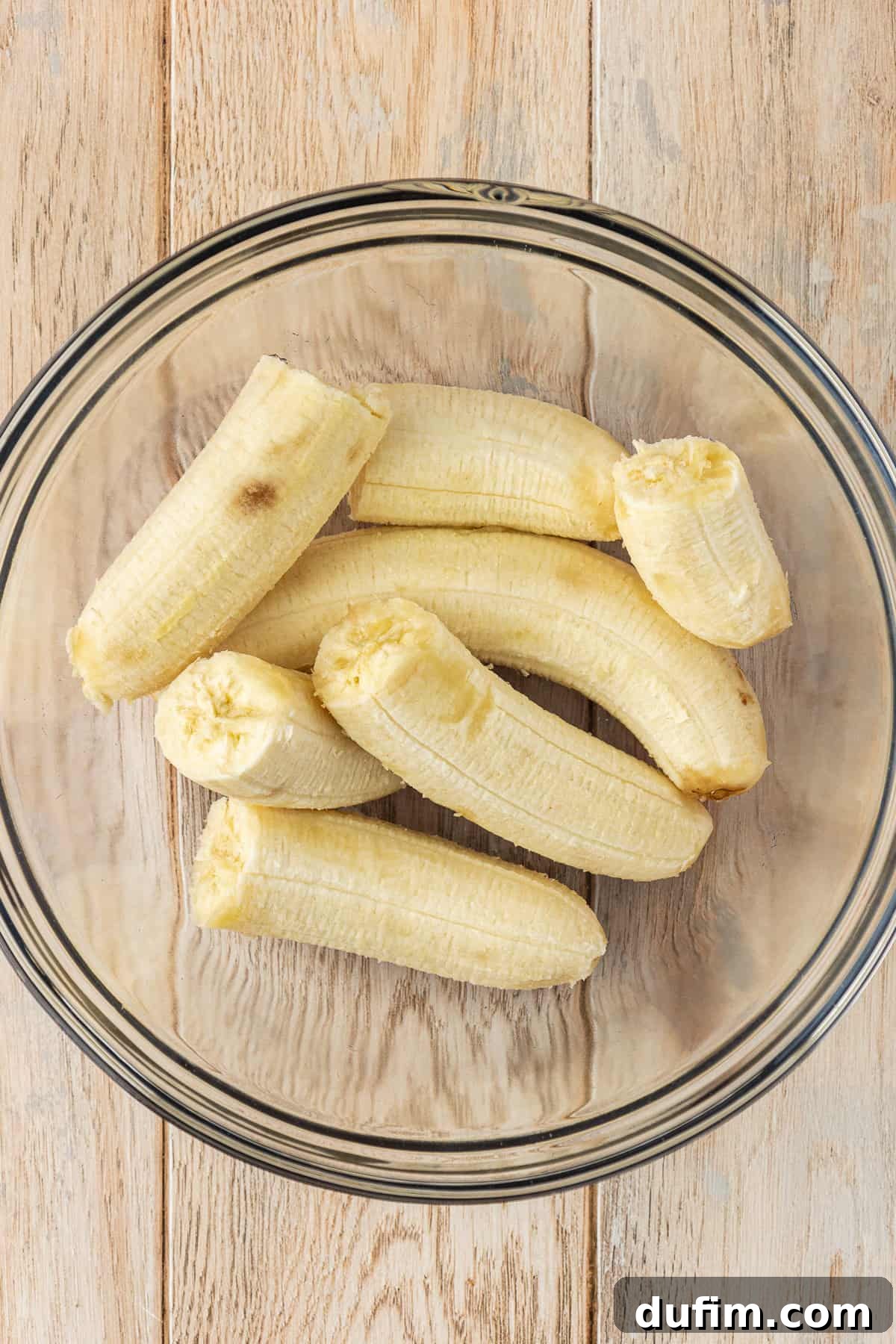 A glass bowl full of banana halves on a wood surface, indicating the start of the preparation process for the banana cake