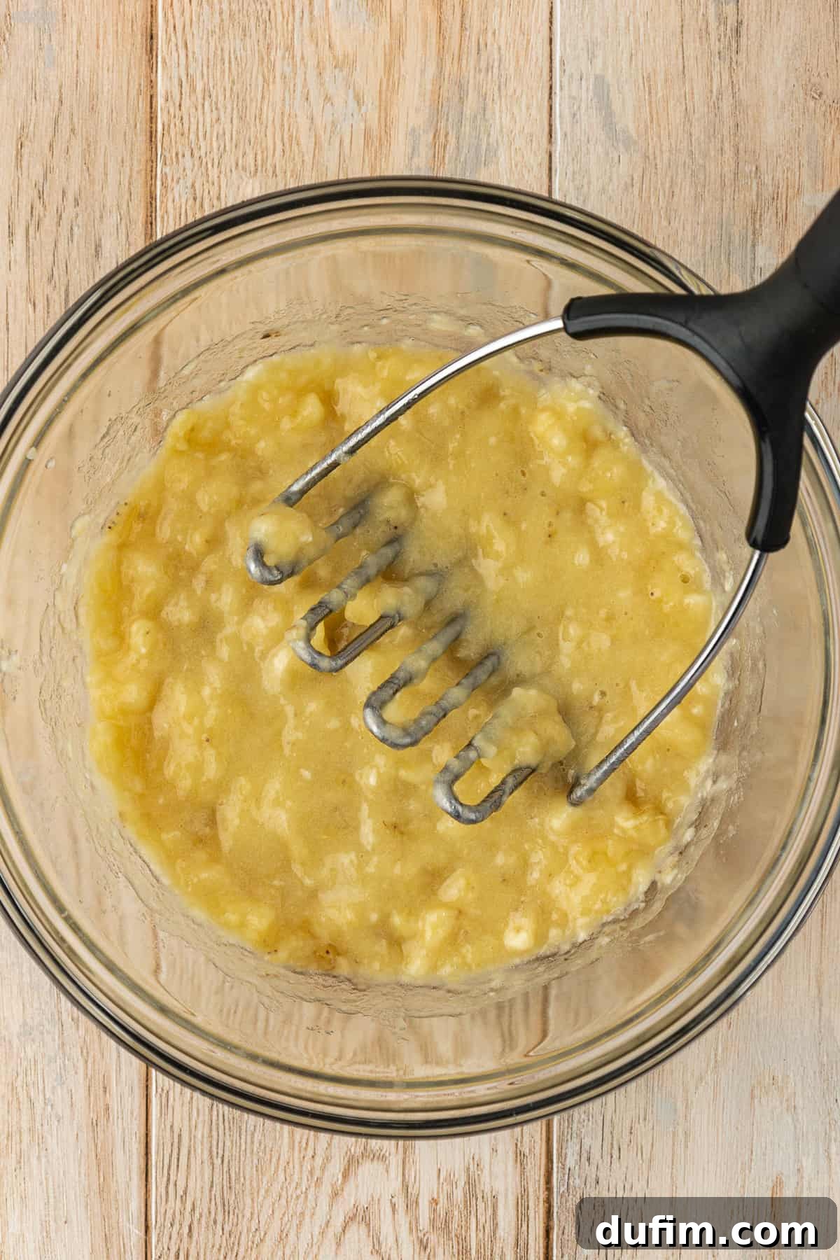 A glass bowl full of mashed banana with a masher resting inside, sitting on a wooden surface, ready to be incorporated into the cake batter