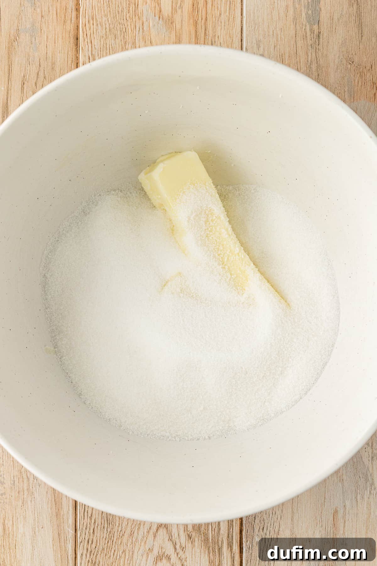 A white bowl on a wood surface containing granulated sugar and a stick of butter, illustrating the initial step of creaming butter and sugar for the banana cake