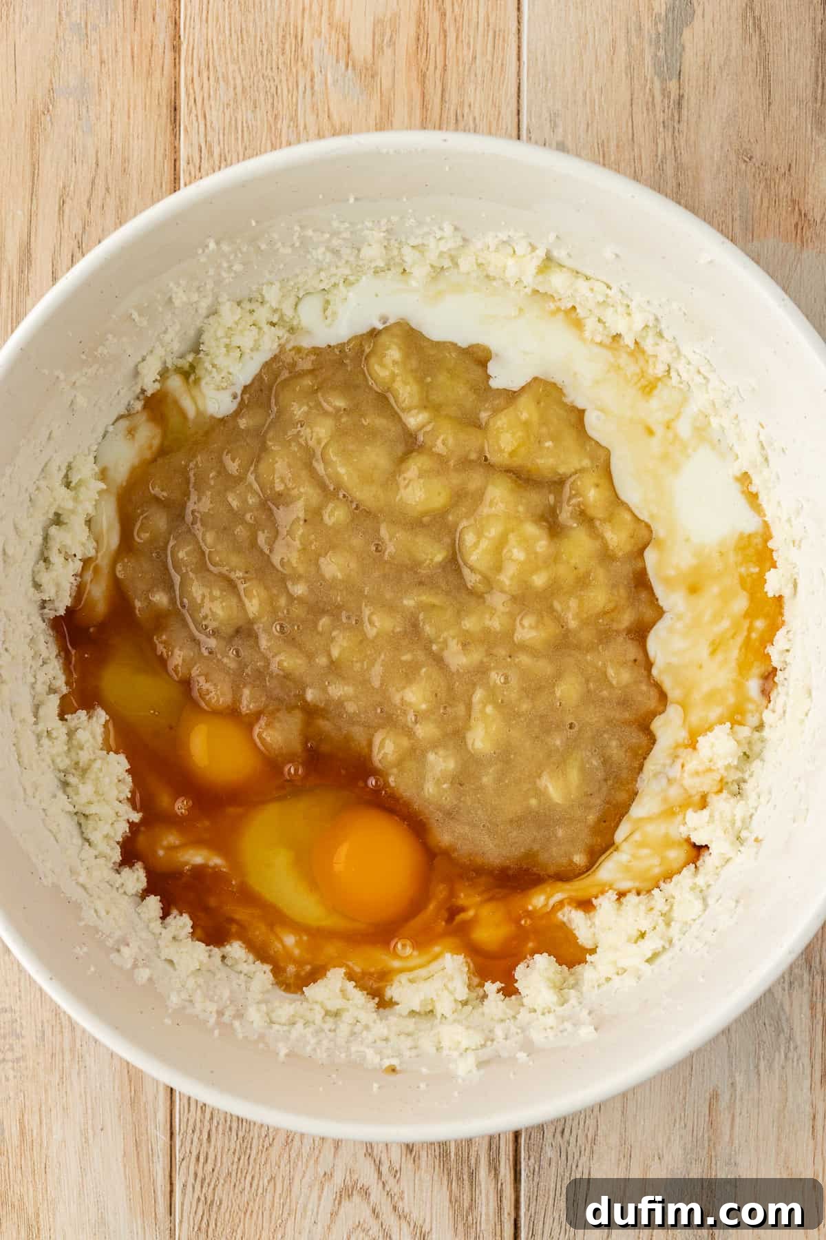A white bowl displaying a butter and sugar mixture, mashed bananas, eggs, buttermilk, and vanilla extract, all unmixed, ready for the next step of combining wet ingredients
