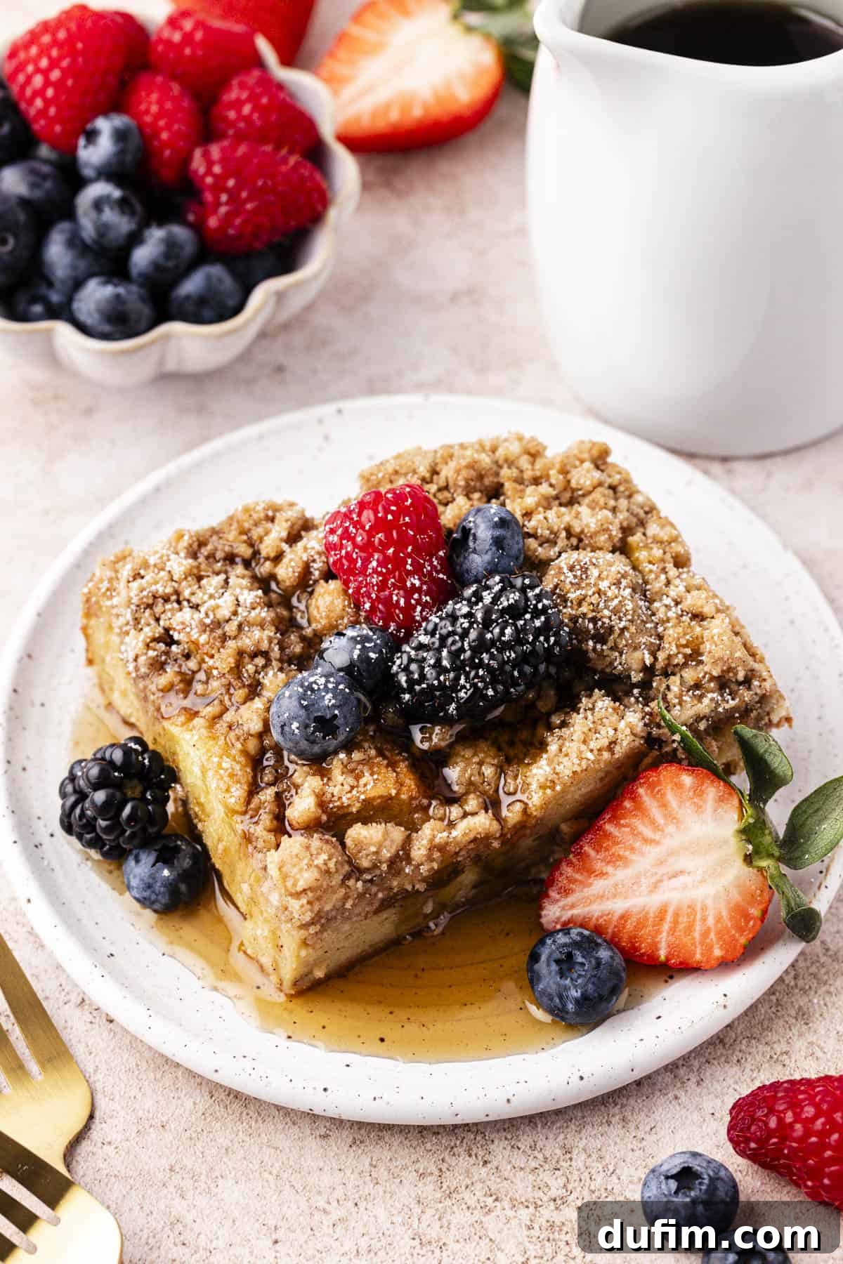 a small plate with a slice of french toast casserole on it topped with maple syrup, fresh berries and a light dusting of powered sugar beside a small bowl of fresh berries, a white dish of syrup, two forks and more berries scattered around