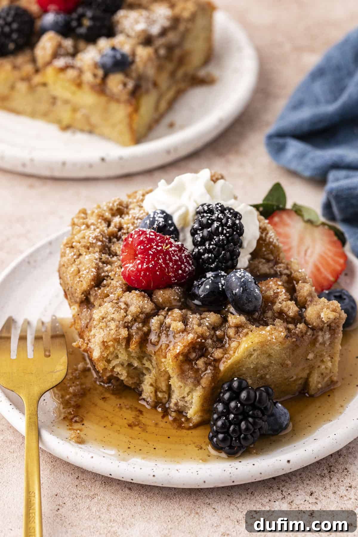 two small plates with a slice of french toast casserole on each, covered in maple syrup, fresh berries, a dollop of whipped cream and a light sprinkle of powdered sugar, with the front plate having a fork leaning on it and a blue kitchen towel in the background