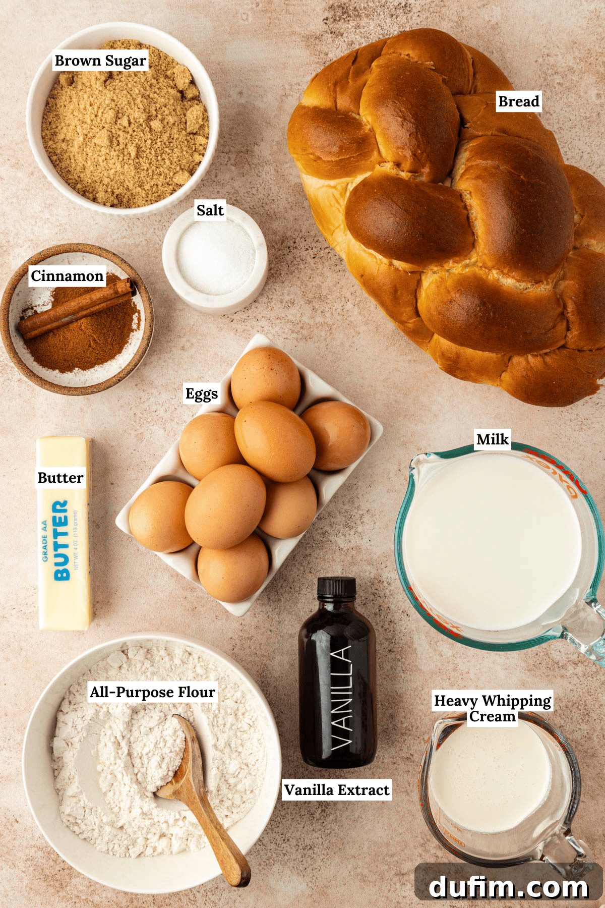 over head view of the ingredients for french toast casserole including a loaf of bread, a bowl of brown sugar, a small bowl of salt, a bowl of cinnamon, eight brown eggs, a stick of butter, a measuring cup of milk, a measuring cup of heavy whipping cream, a bottle of vanilla extract and a bowl of all-purpose flour
