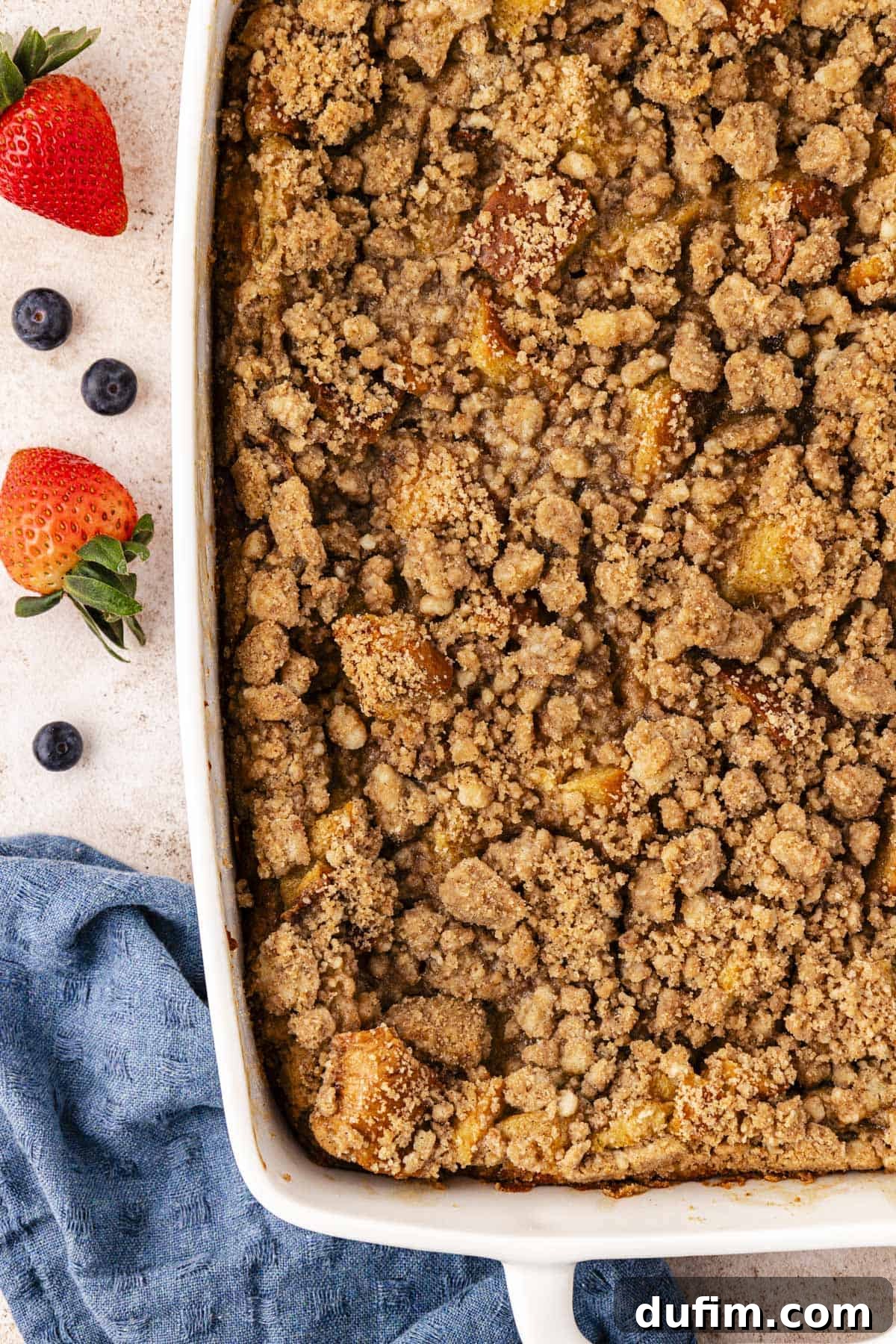 a large white baking dish of french toast casserole with crumb topping beside a blue kitchen towel and fresh blueberries and strawberries scattered around