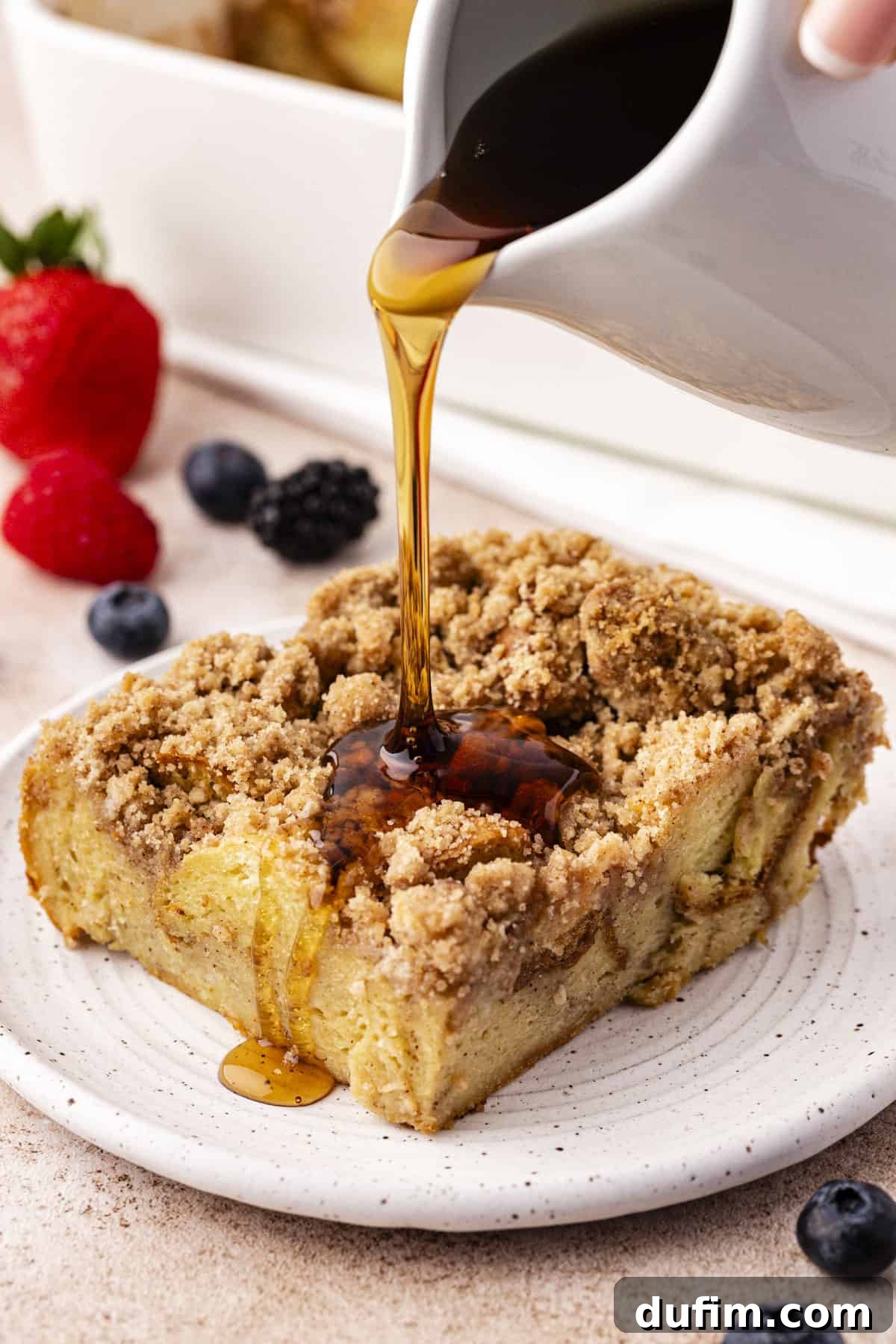 a slice of french toast casserole on a small plate with maple syrup being poured over the top and fresh berries scattered around