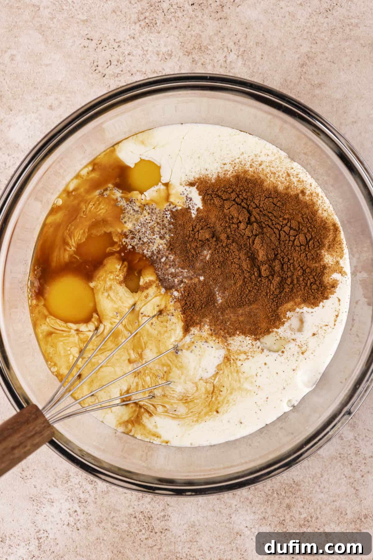 a clear glass bowl with eggs, whole milk, heavy cream, brown sugar, cinnamon, salt and vanilla extract in it with a wire whisk leaning inside the bowl 