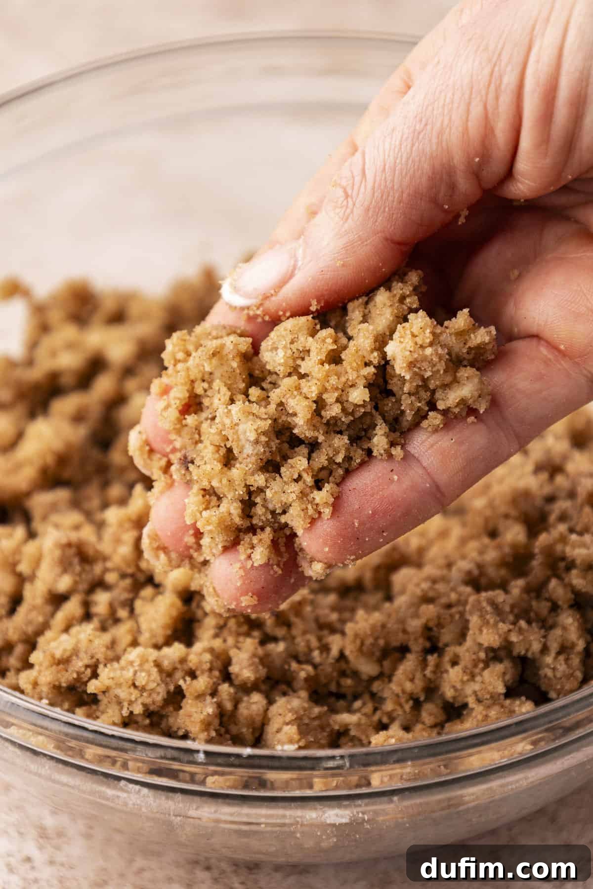 a hand holding crumb topping over top of a clear glass bowl full of more crumb topping