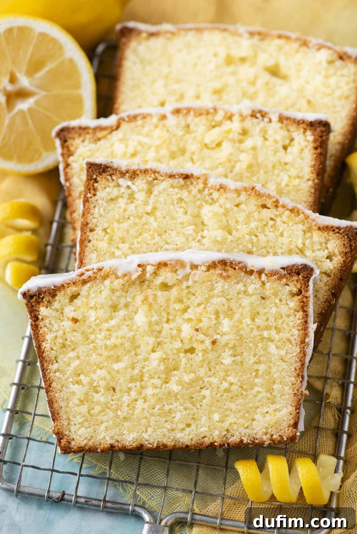 Slices of glistening lemon pound cake on a wire rack, surrounded by fresh lemon slices and aromatic lemon peels.