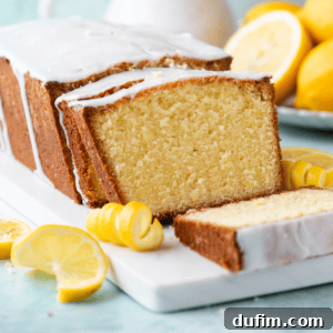 A sliced loaf of lemon pound cake on a white cutting board on a light blue surface, adorned with fresh lemon slices and peels, and more lemons in the background.