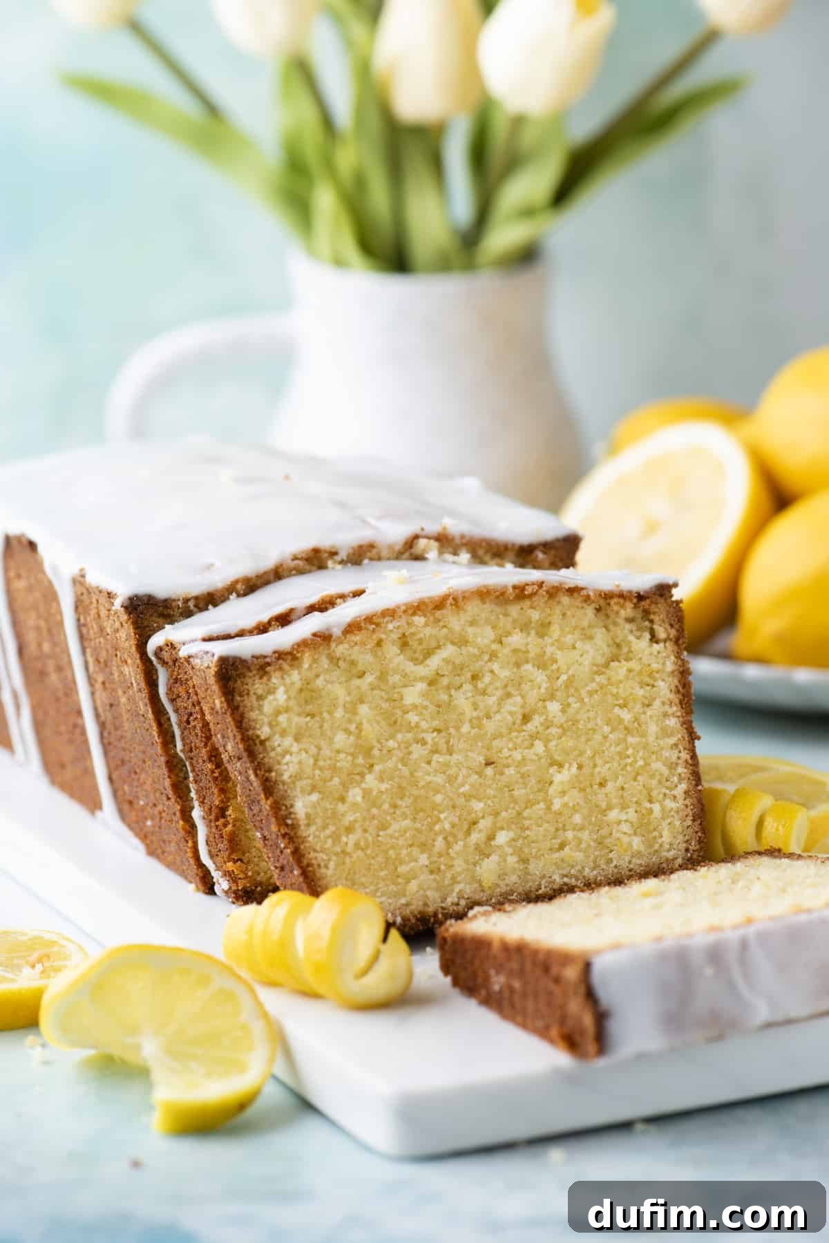 Sliced lemon pound cake resting on a pristine white cutting board, artfully arranged with fresh lemon slices, delicate peels, and soft flowers in the background.