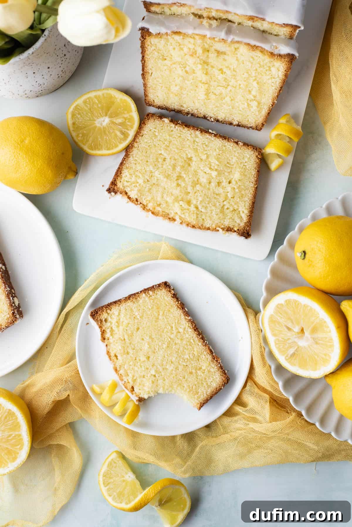 An inviting overhead shot of a lemon pound cake, half-sliced on a white cutting board, surrounded by small white plates with individual cake slices, whole lemons, and vibrant fresh flowers.