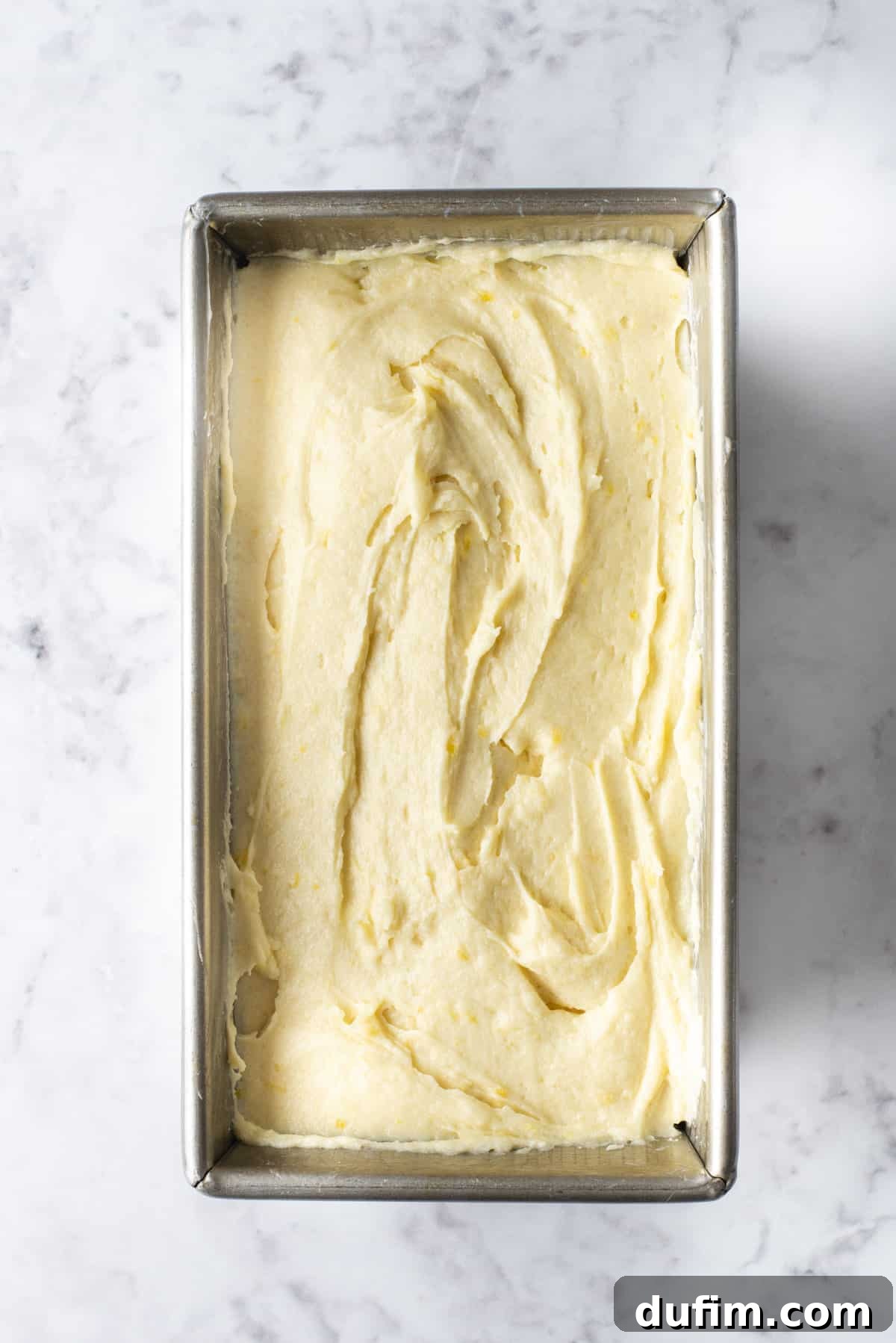 Thick lemon pound cake batter perfectly settled in a loaf pan, ready for baking on a clean countertop.