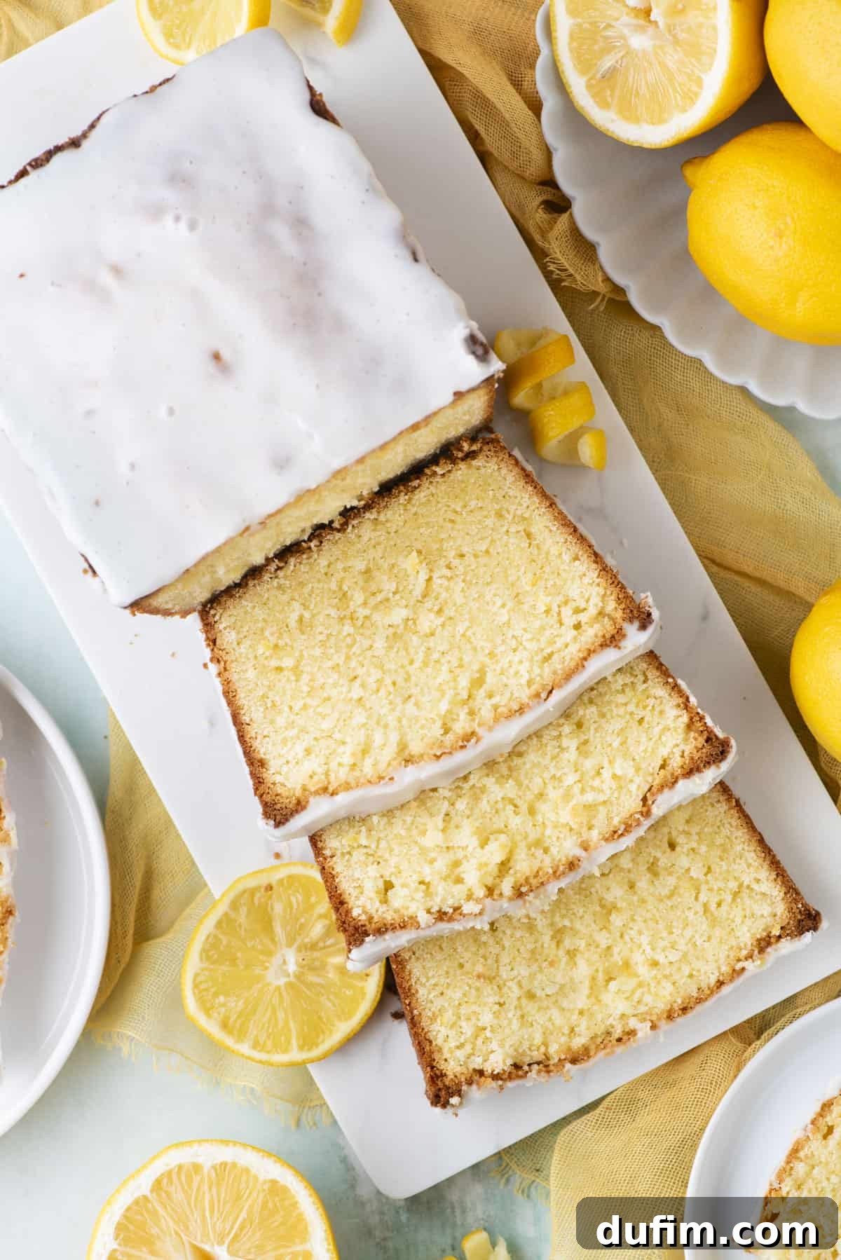 An overhead shot of a baked lemon pound cake, half-sliced, on a white cutting board. It's surrounded by a bowl of fresh whole lemons, lemon slices, and loose peels, creating a rustic and inviting scene.