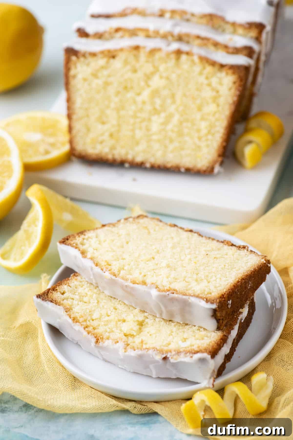 Two enticing slices of lemon pound cake, perfectly glazed, served on a small white plate. The background features more of the cake, along with fresh lemon slices and peels.