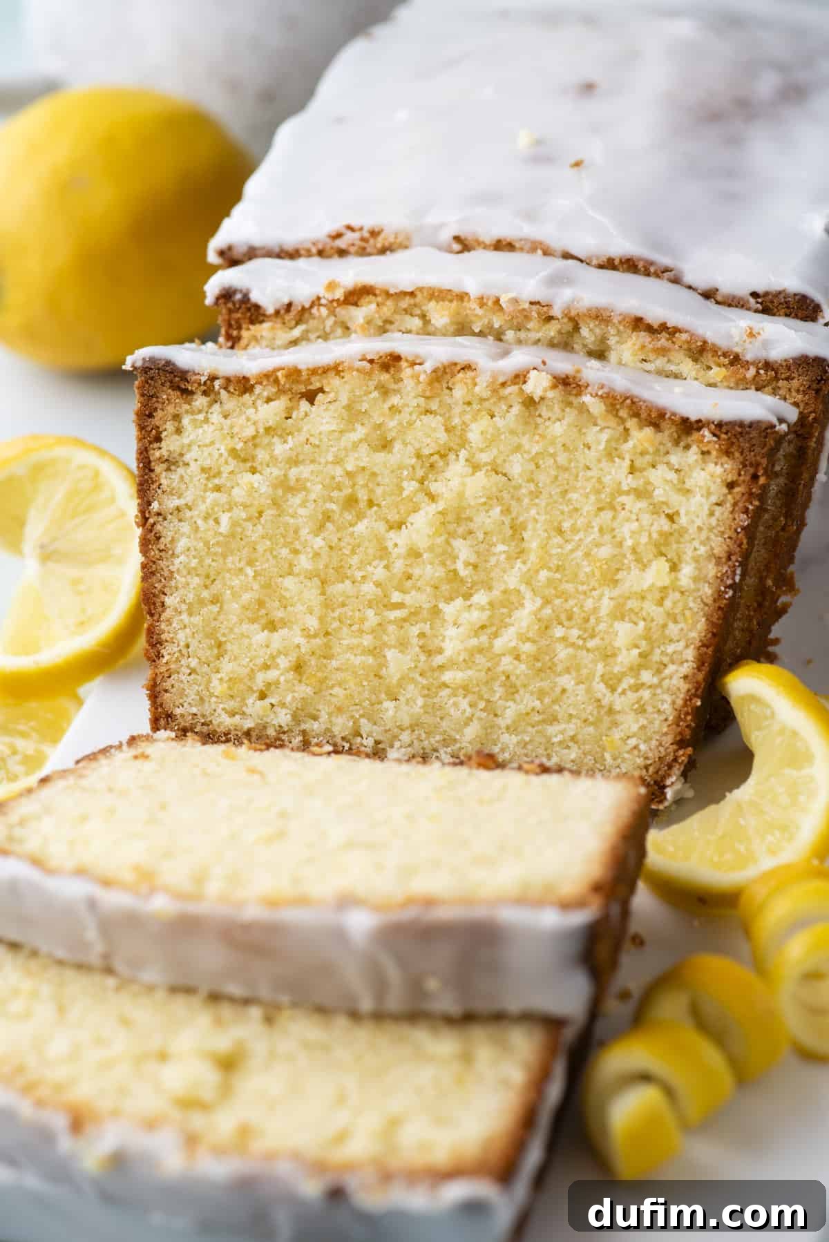 A beautifully sliced lemon pound cake, glistening with glaze, surrounded by scattered fresh lemon slices and aromatic lemon peels.