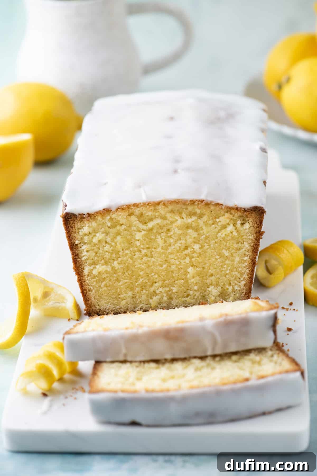 A glorious sliced lemon pound cake on a white cutting board, elegantly framed by fresh lemon slices and vibrant lemon peels.