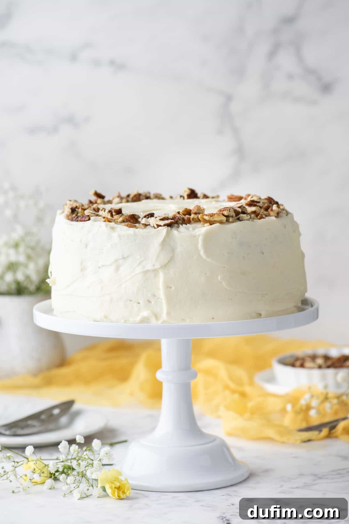 A beautifully frosted hummingbird cake topped with chopped pecans on a white cake stand, surrounded by elegant white and yellow flowers, a yellow linen, and a bowl of pecans in the background, showcasing its delightful presentation.