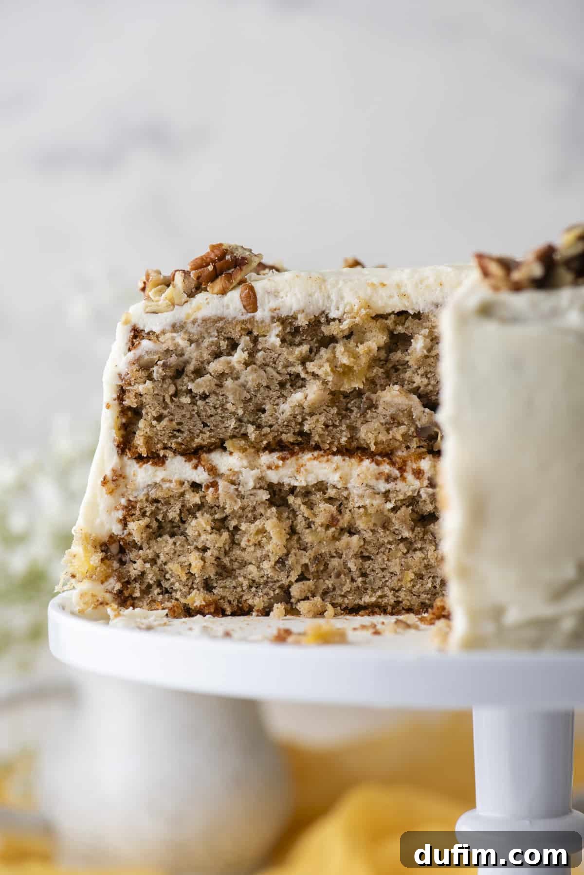 A close-up, side view of a two-layer hummingbird cake on a white cake stand, with one slice already removed. This view clearly exposes the moist internal structure of the cake layers and the generous, creamy frosting in between, highlighting its inviting texture.