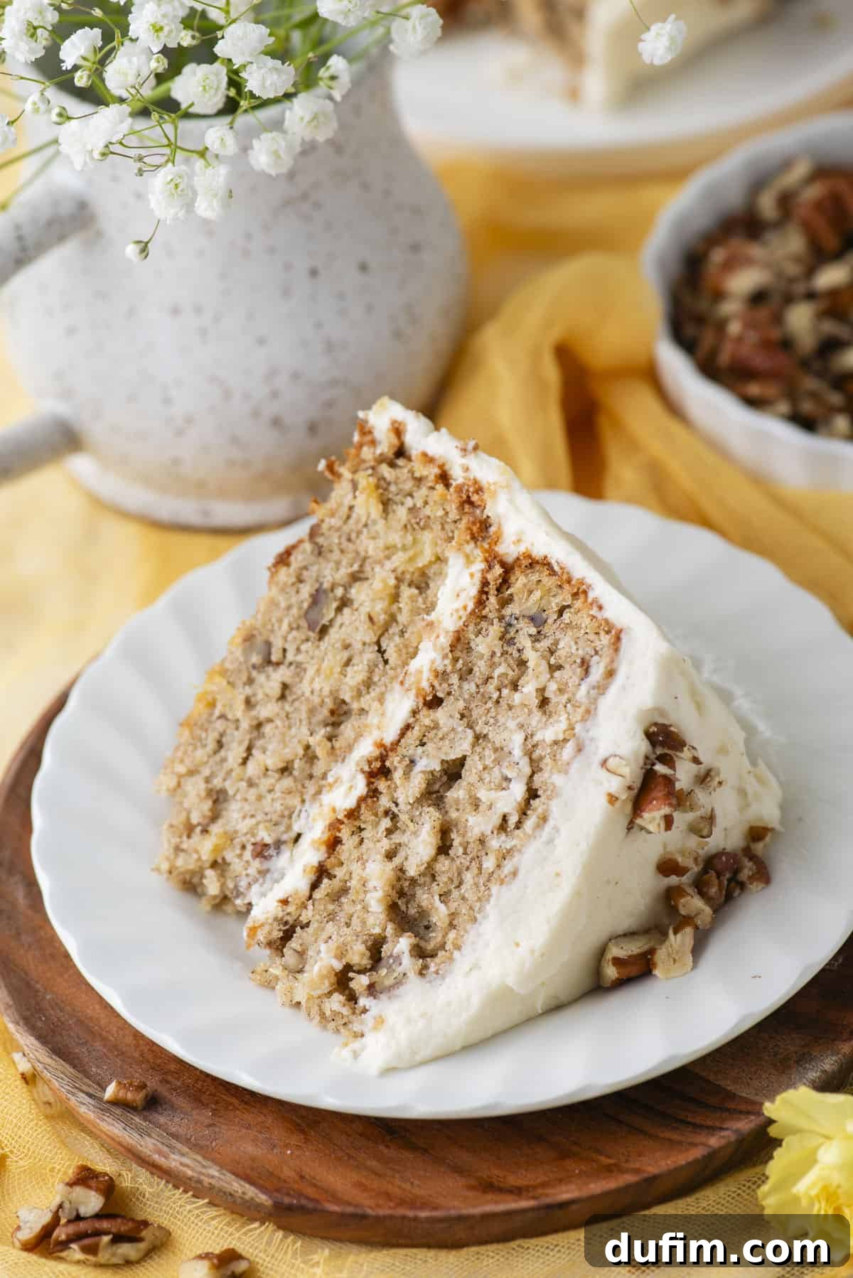 A pristine slice of hummingbird cake on a small white plate, beautifully presented on a round wooden base. Beside it, a small dish of pecans and an elegant vase of white flowers complete the inviting composition, emphasizing the cake's delicious appeal.