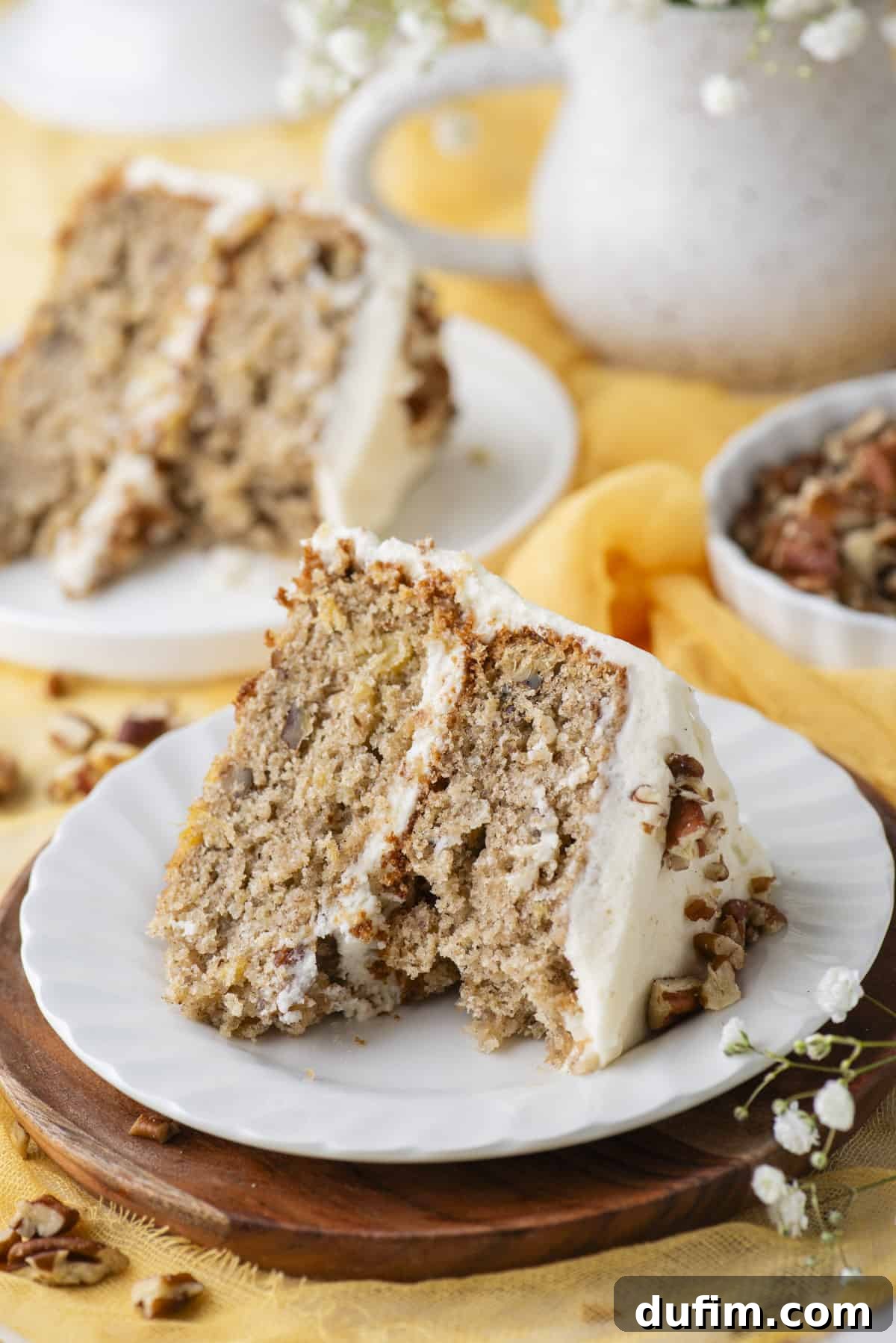 A perfectly sliced piece of hummingbird cake on a white plate, resting on a wooden coaster. One bite is missing from the slice, revealing the moist layers and frosting. Chopped pecans are scattered around, with white flowers and another slice of cake in the soft background, highlighting its delectable texture.
