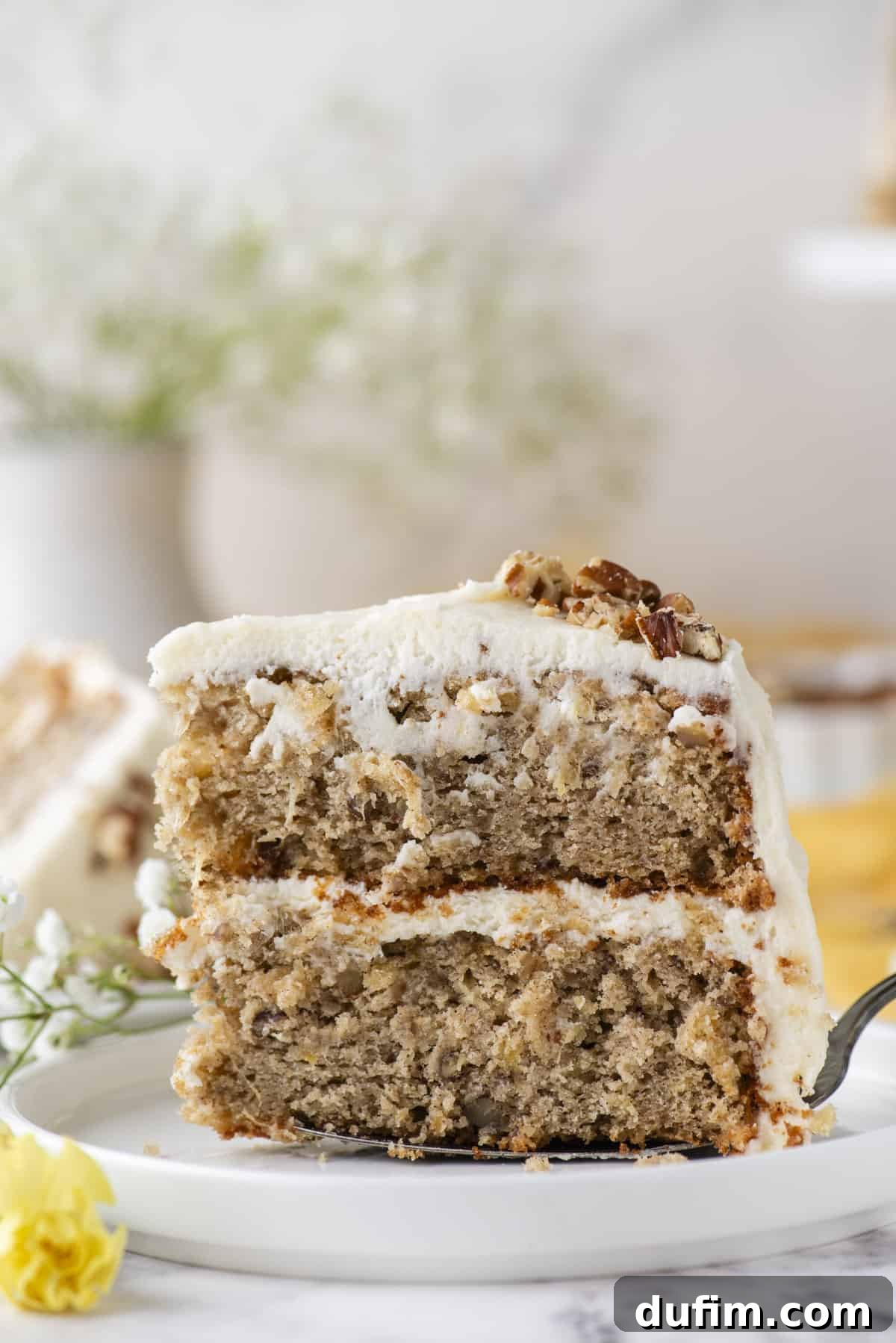 A slice of two-layer hummingbird cake being carefully transferred onto a white plate with a spatula, beautifully showcasing the distinct layers of cake and the thick cream cheese frosting in between. Elegant white and yellow flowers are visible on the left, adding to the aesthetic appeal.