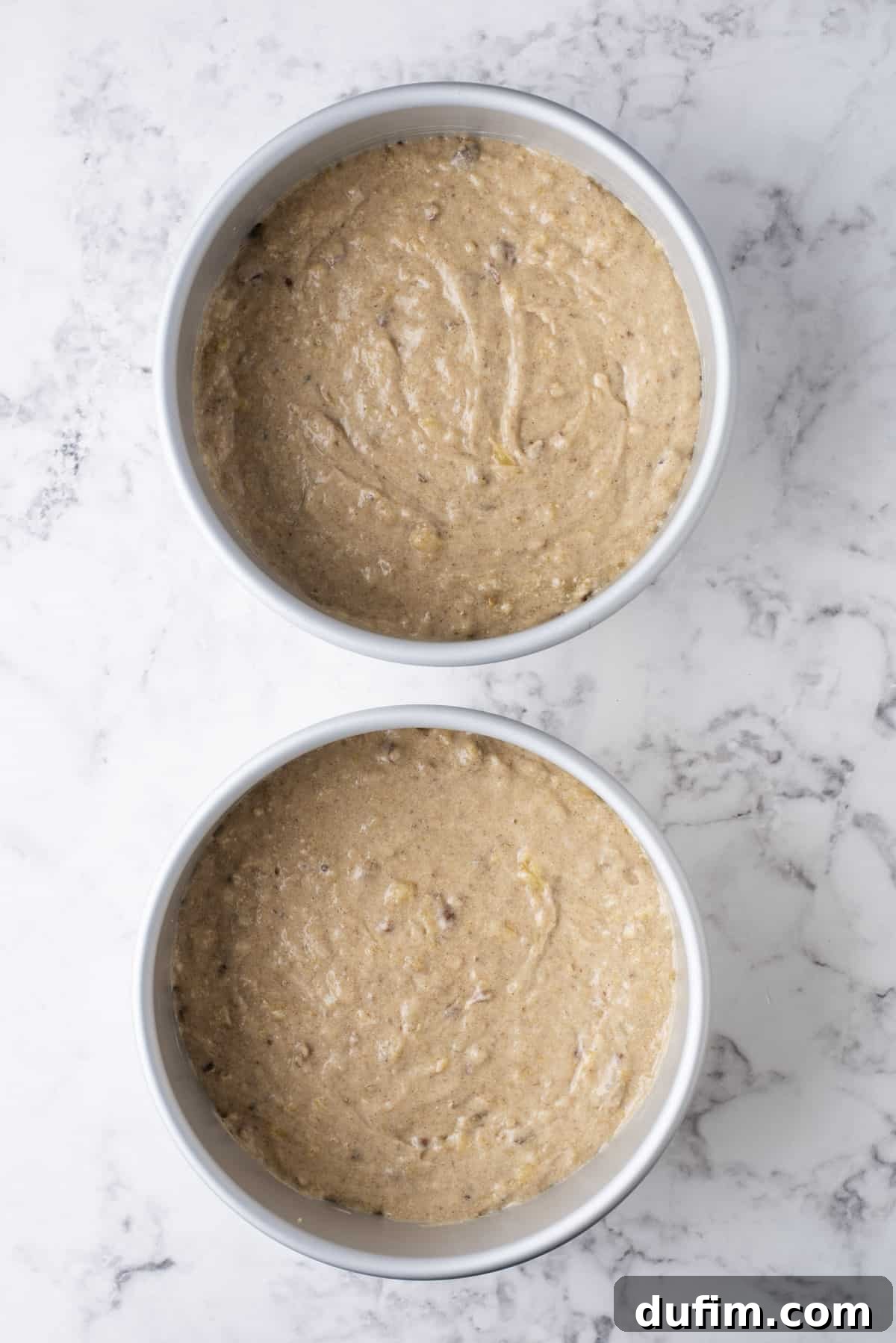 Two round cake pans filled with a rich, lumpy hummingbird cake batter, ready for baking. The batter shows visible pieces of fruit and nuts.