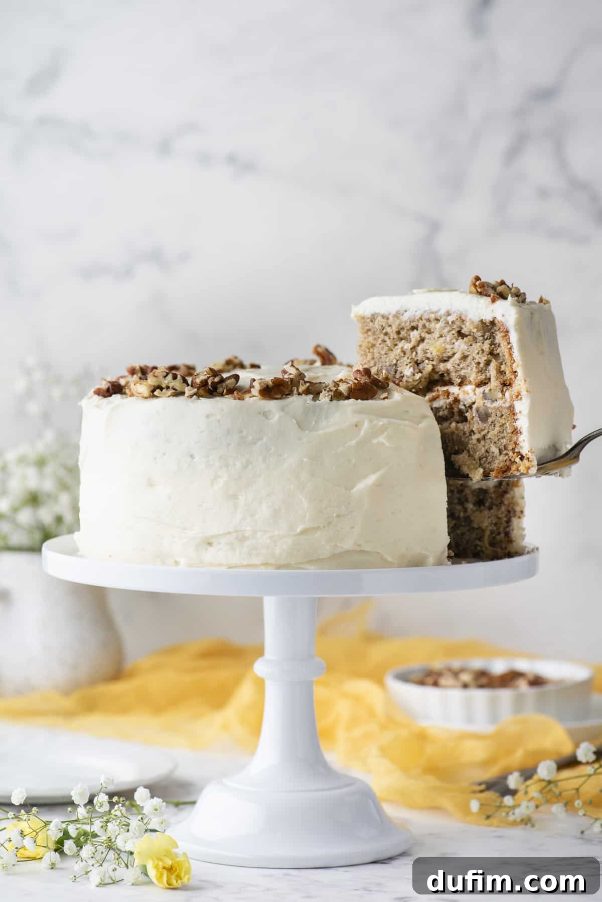 A beautifully decorated hummingbird cake on a white cake stand, with one slice being lifted away with a spatula, showcasing the layers of cake and frosting. The background features white and yellow flowers, a yellow cloth, and a white dish of pecans, creating an inviting scene.
