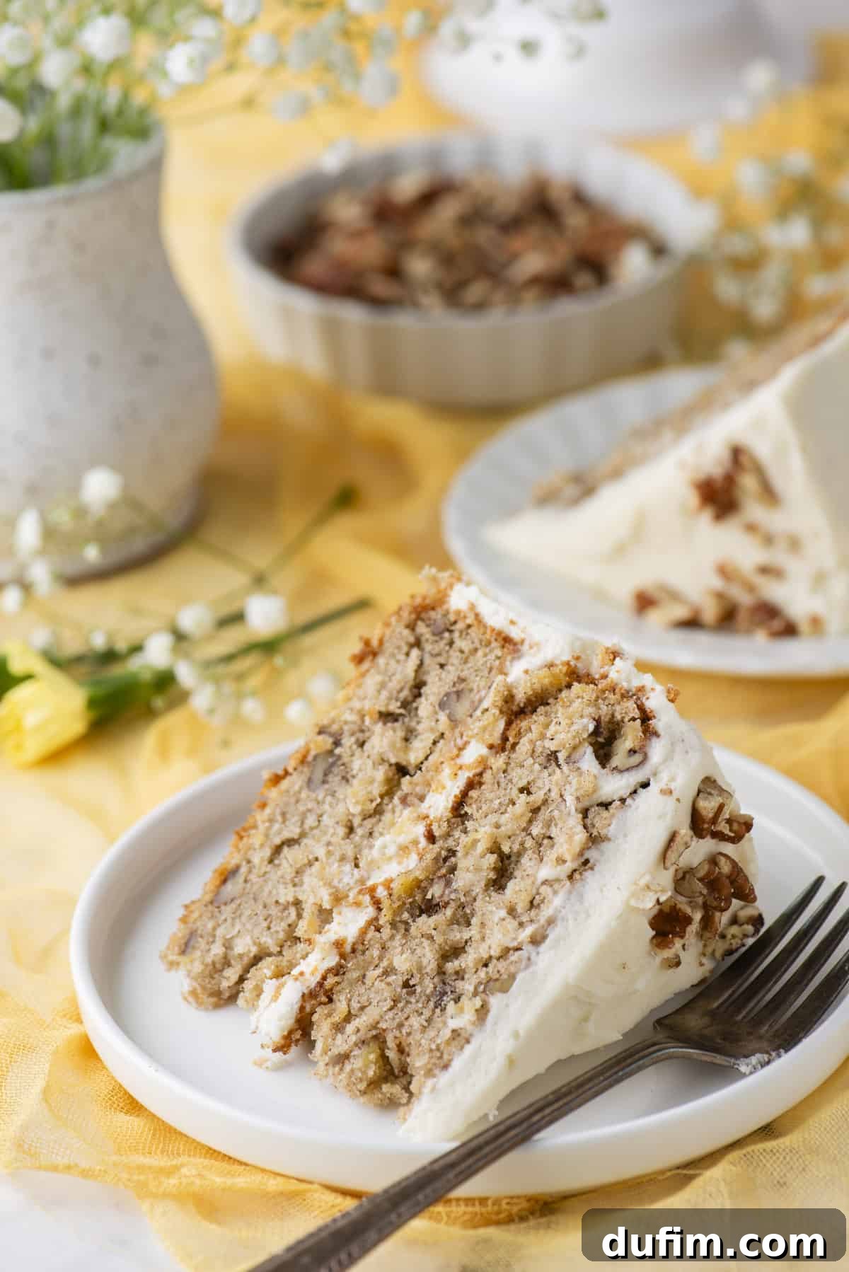 A delightful slice of two-layer hummingbird cake on a small white plate, with a fork resting beside it, positioned on a yellow linen. In the background, there are yellow and white flowers, another slice of cake on a plate, and a dish of pecans, creating a warm and inviting atmosphere.