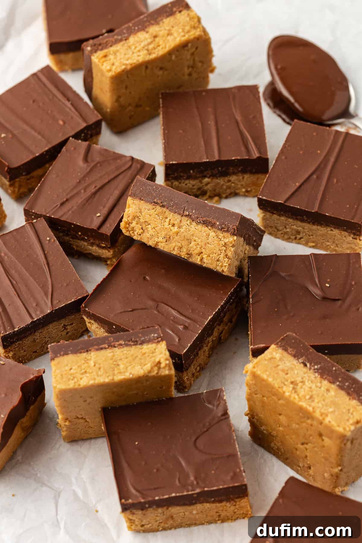 Peanut butter bars scattered around on white parchment paper, some on their sides, some flat, some leaning on others, and one spoon of melted chocolate beside them, highlighting the rich chocolate topping and creamy peanut butter base.