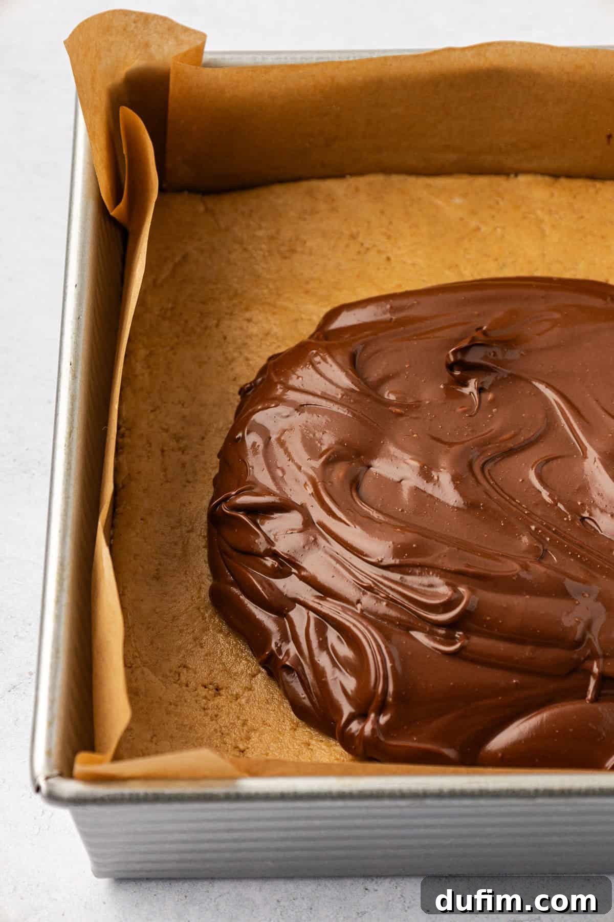 Close up of a square pan lined with brown parchment paper with a smooth peanut butter layer topped with a large dollop of a melted chocolate mixture, beginning to be spread.