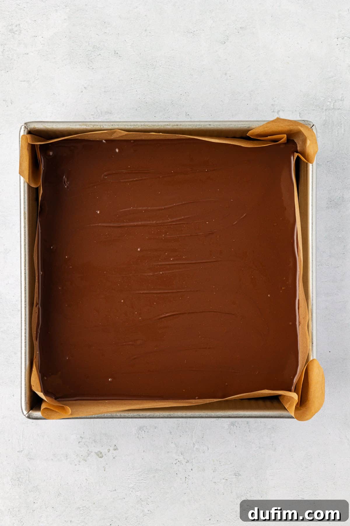 Overhead view of a square pan lined with parchment paper with a smooth, even chocolate layer completely covering the peanut butter base.