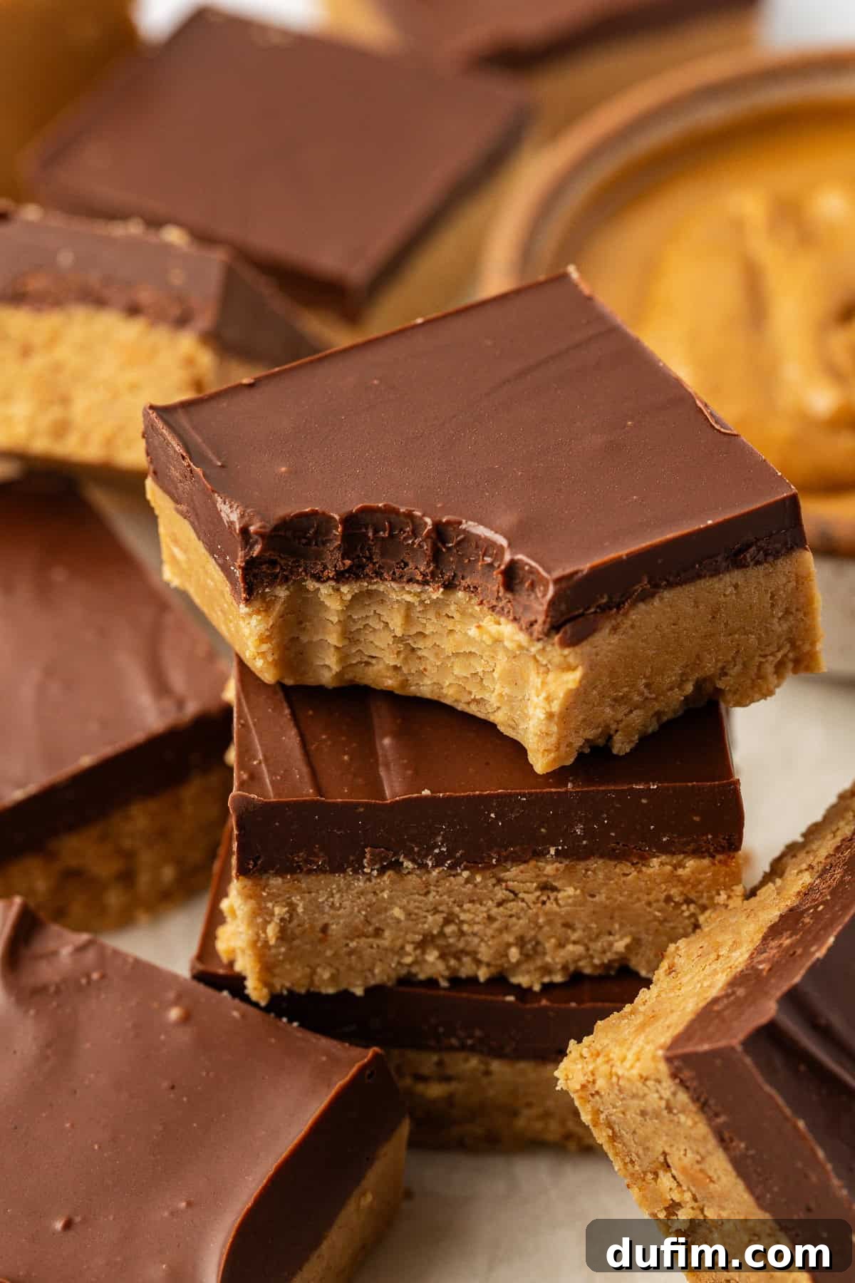 A stack of three peanut butter bars with a bite taken out of the top bar, revealing the creamy layers. More peanut butter bars are scattered casually around on parchment paper.