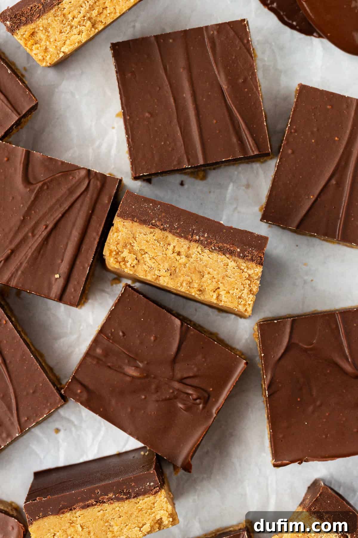 Peanut butter bars scattered all around on white parchment paper, some laying flat and some on their sides showing the distinct layers of peanut butter and chocolate.