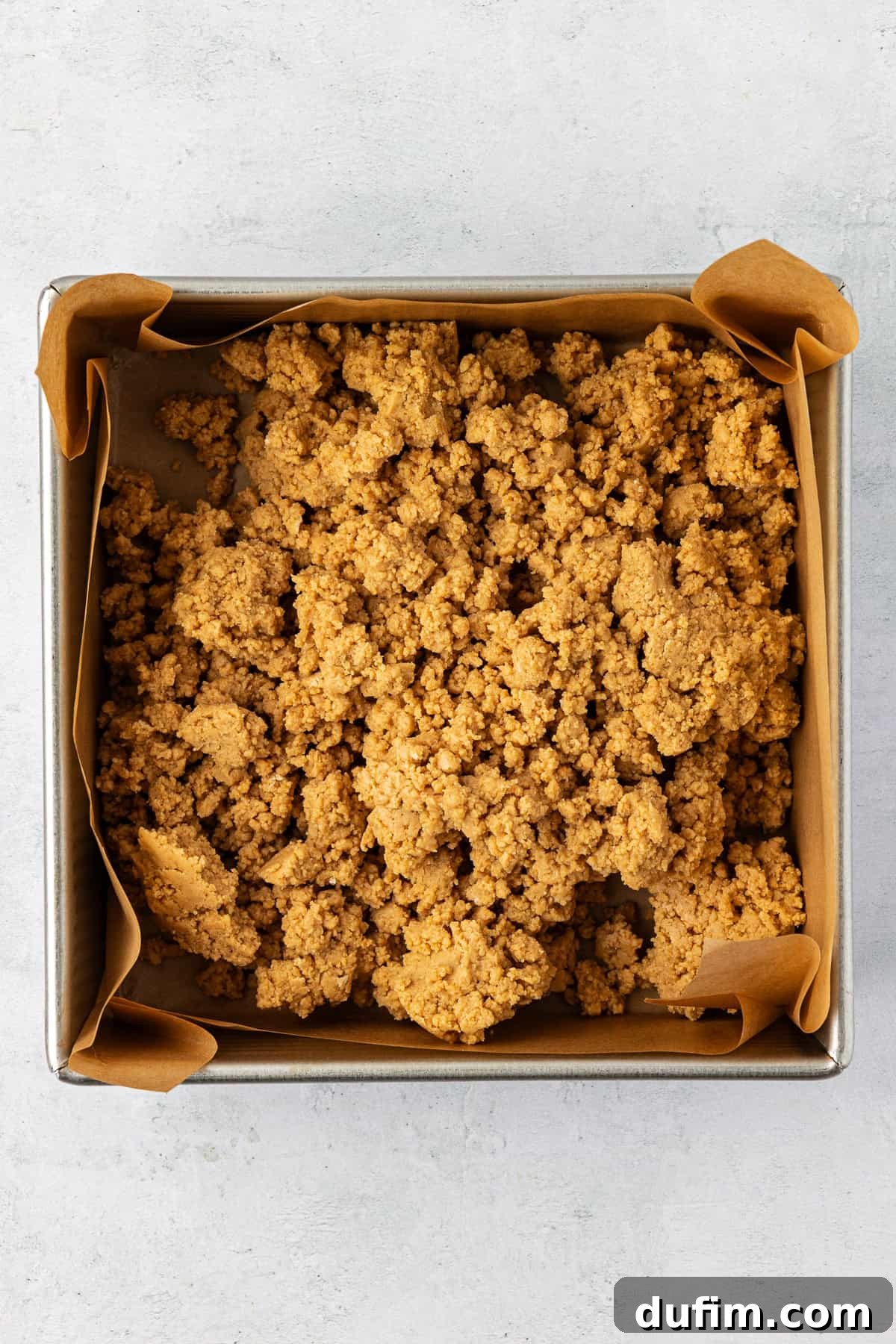 A square pan lined with brown parchment paper with a peanut butter and graham cracker mixture dolloped inside of it, ready to be pressed down.