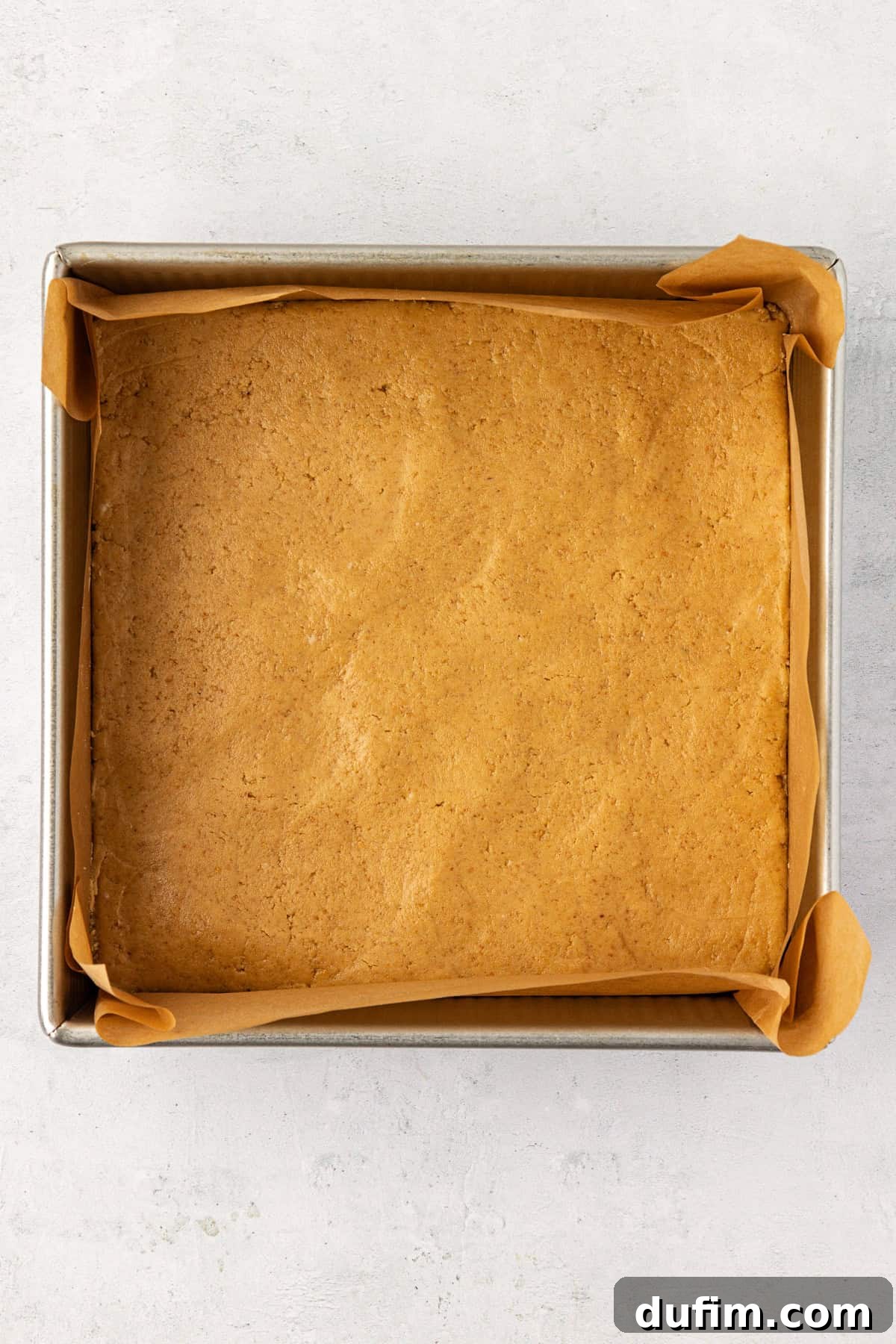 A square pan lined with parchment paper with a smooth, compact layer of peanut butter mixture pressed evenly into the bottom.