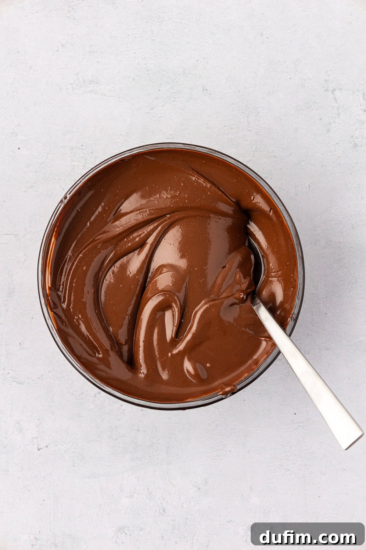 A melted chocolate and peanut butter mixture in a clear glass bowl with a spoon stuck in it, showing its luscious, smooth texture.