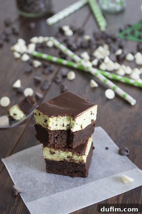 A close-up shot of a perfectly layered Mint Chip Mousse Brownie slice, showcasing the distinct layers: a rich, dark chocolate brownie base, a thick, fluffy green mint mousse, and a thin, shiny chocolate glaze on top. White and dark chocolate chips are scattered around, adding a decorative touch.