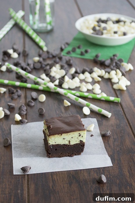 A tray of freshly baked brownies, topped with a smooth layer of vibrant green mint mousse, is seen in the foreground. A bowl of melted chocolate sits nearby, ready to be transformed into the final glossy glaze. The image captures the multi-step process of creating this layered dessert.