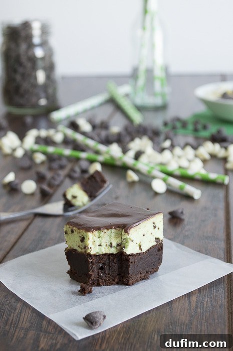 A stack of perfectly cut Mint Chip Mousse Brownies on a piece of parchment paper, adorned with mint leaves and chocolate chips. The distinct layers are clearly visible, highlighting the dessert's elegance and craftsmanship.